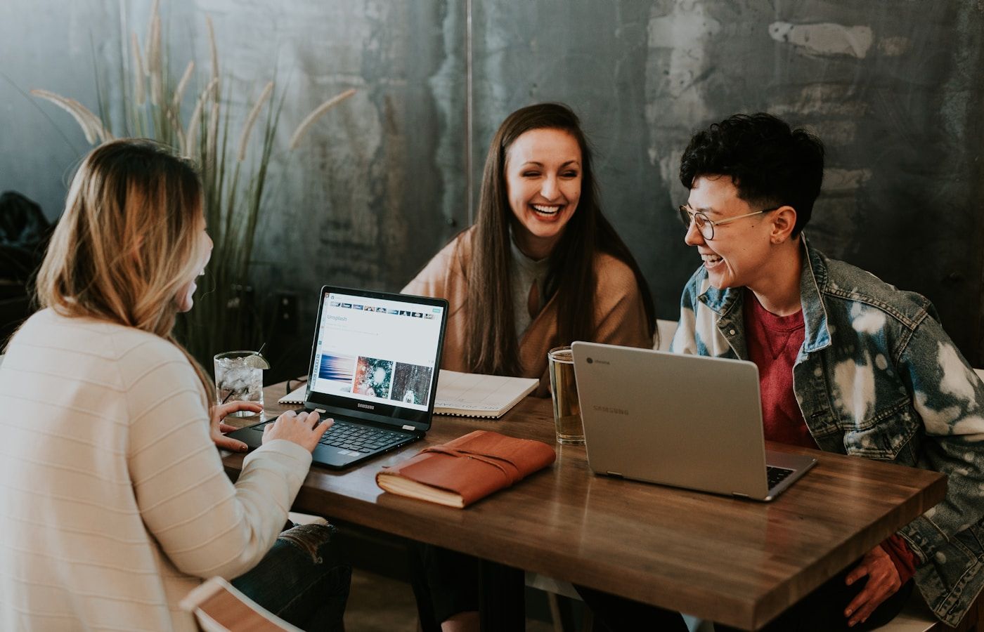 Three people laughing together around a table with laptops during a friendly working session