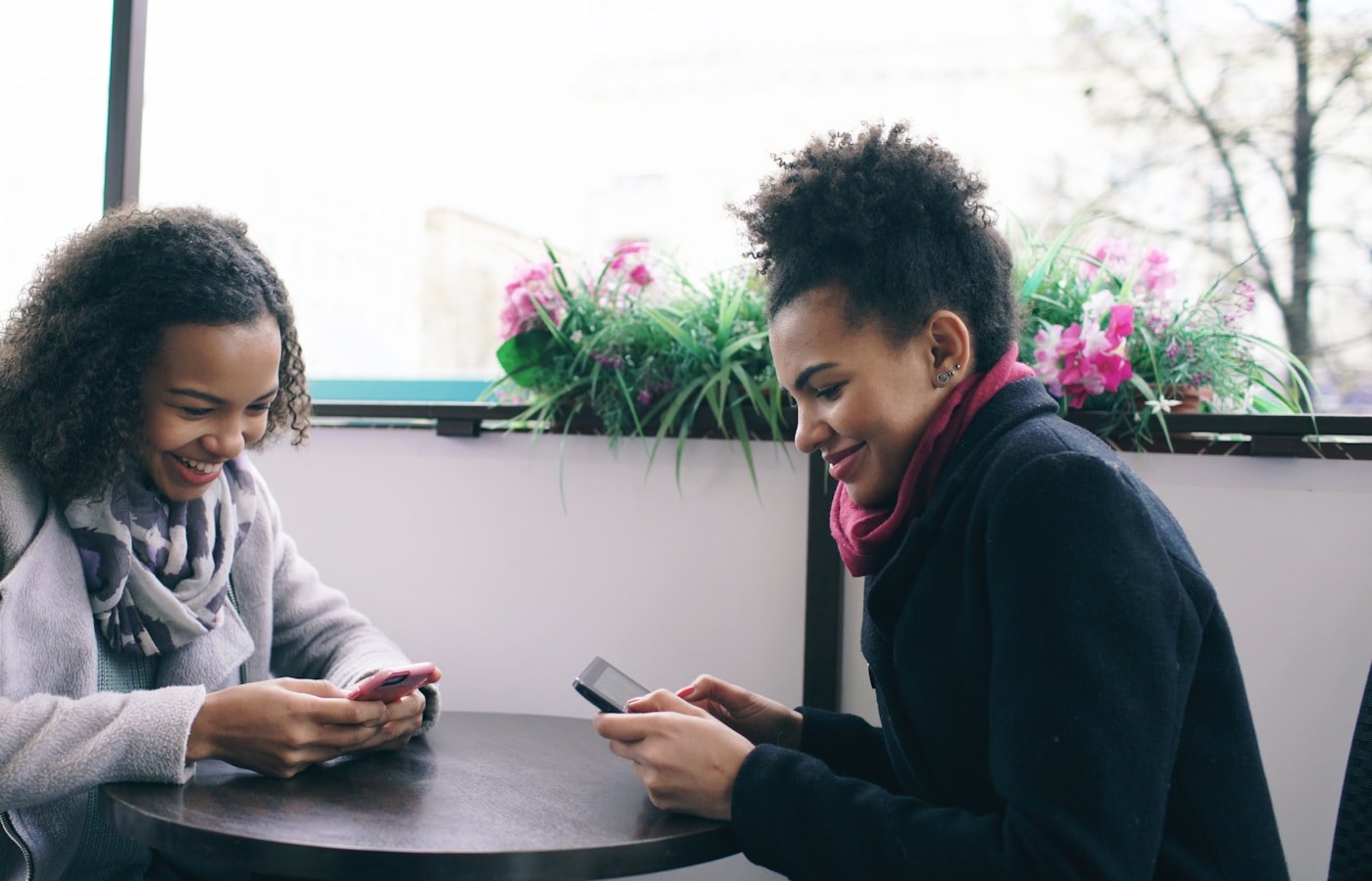 Two people sitting together at a cafe table using their phones and smiling